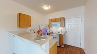 Located at 4510 Tesla Park Dr in Wilmington, NC, this single-floor unit offers a practical and well-organized layout designed for comfortable everyday living. The kitchen is a functional highlight, featuring warm honey-toned cabinetry, white appliances including a coil-burner range, over-the-range microwave, and refrigerator. A peninsula breakfast bar with a light countertop extends into the living area, creating an open, connected feel between the kitchen and main living space. The kitchen is finished with durable hardwood-style flooring that adds warmth and is easy to maintain. The living area is generously proportioned, carpeted in neutral tones, and fitted with a ceiling fan for year-round comfort. A door with a transom window allows natural light to fill the space, adding brightness to the open floor plan. An additional carpeted room with its own ceiling fan serves well as a bedroom or flex space, offering a clean, versatile canvas. The bathroom is neatly appointed with a single-sink vanity featuring light wood cabinetry, a wall mirror with overhead lighting, and a shower/tub combination. Throughout the unit, the bright white walls and consistent neutral palette create a clean, cohesive ambiance.