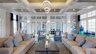 This elegantly appointed single-floor space showcases a refined coastal-inspired aesthetic that blends classic architectural detail with contemporary furnishings. The grand entrance foyer welcomes visitors through floor-to-ceiling French doors topped with an arched transom window, flooding the space with natural light and offering views of the outdoor surroundings. Rich hardwood flooring runs throughout, anchoring the open, airy layout.

The centerpiece of the space is a generously proportioned lounge area featuring dramatic coffered ceilings with wood-paneled insets and recessed lighting, complemented by a striking double-ring modern chandelier. Crisp white columns and extensive glass-paned interior French doors with transom windows divide the space while maintaining a sense of openness and flow between areas.

The seating arrangement consists of plush neutral sofas and tailored wingback chairs in coordinated blue-gray tones, set atop a patterned area rug. Decorative accents in navy, teal, and aqua add vibrant coastal color throughout. Wall sconces and table lamps provide warm ambient lighting alongside the abundant natural light streaming in from large exterior windows dressed with bold navy and teal drapery panels.

Adjacent rooms, visible through the interior glass partitions, suggest additional lounge and activity areas, contributing to the expansive, multi-functional character of the overall space.