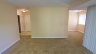 This single-floor space presents a clean, straightforward layout designed for comfortable living. The main living area is finished with wall-to-wall carpeting in a neutral beige tone, providing a warm and cohesive foundation throughout the space. The walls are painted in a soft, light cream color that enhances the sense of openness and reflects available light effectively.

A partial dividing wall creates a subtle separation between areas of the space while maintaining an open feel, allowing light to flow through naturally. Flush-mounted ceiling lighting provides functional illumination in the main area, while additional track lighting is visible in the adjacent section, offering more focused lighting options.

Toward the back of the space, a hallway leads to additional rooms, featuring a mirrored sliding closet door that adds depth and reflects light. A solid entry door with standard hardware is also visible from this vantage point, offering a clear sense of the unit's layout and flow.

The overall ambiance is neutral and move-in ready, with a simple, unfussy design that provides a versatile canvas for a variety of interior styles and furnishing arrangements.