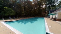 This single-story community clubhouse is designed to offer both functionality and comfort in a well-maintained setting. The exterior showcases classic architecture with white vinyl siding, brick accents, black shutters, and a pitched shingled roof with dormer windows — lending the building a timeless, residential character. A paved pool deck leads directly to the building's rear entrance, creating a seamless indoor-outdoor connection.

The main entry features a glass-paneled door flanked by sidelights that frame views of the surrounding parking area and landscaped grounds. Step inside to a bright, open lobby finished with large-format tile flooring, beadboard wainscoting, and soaring ceilings that allow natural light to fill the space. Framed artwork and a console table add welcoming touches near the rear glass doors, which look directly out onto the pool area.

A carpeted hallway extends from the lobby, lined with framed artwork and trimmed with chair-rail molding, guiding visitors deeper into the facility's interior spaces.

The centerpiece of the outdoor area is a generously sized rectangular swimming pool with clear turquoise water, surrounded by a concrete deck furnished with lounge chairs. Mature trees enclose the pool area, providing natural shade and a serene, private atmosphere — making it an ideal setting for relaxation and recreation.