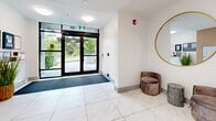 Located at 2590 Lonsdale Avenue in North Vancouver, BC, this building lobby offers a polished and welcoming first impression. The hallway is finished with large-format marble-effect porcelain floor tiles that lend a clean, upscale aesthetic throughout the space. Automatic glass entry doors provide easy access from Lonsdale Avenue, framing views of the lush greenery outside and allowing natural light to flood the interior.

The walls are finished in crisp white, accented by warm wood slat paneling that adds texture and a contemporary touch. A decorative round gold-framed mirror and a plush velvet accent chair provide an inviting focal point near the entrance. An abstract wall painting further enhances the curated, design-forward atmosphere.

A dedicated mailroom area features a full bank of individual resident mailboxes, neatly integrated into the warm wood slat wall, keeping the space organized and functional. Resident notices and building information are clearly displayed near the entry, ensuring easy communication.

Flush ceiling light fixtures provide consistent, even illumination throughout. The lobby connects directly to the main interior corridor, with elevator access visible from within. Thoughtful details — from the decorative greenery to the carefully selected furnishings — make this lobby a refined and practical gateway into the building.