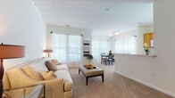 This well-appointed single-floor residence offers a thoughtfully designed layout that balances comfort and functionality across its key living spaces. The open-concept living and dining area serves as the heart of the home, bathed in abundant natural light streaming through multiple large windows dressed with sheer white curtains and blinds. Plush neutral carpeting, crisp white walls, and soaring ceilings contribute to an airy, welcoming atmosphere throughout the main living zone. The dining area, situated adjacent to the living room, features its own generous window arrangement, making it a bright and inviting space for everyday meals. The galley-style kitchen is outfitted with warm honey-toned wood cabinetry, white countertops, and hardwood-style flooring, offering ample storage and prep space in an efficient layout. Two well-sized bedrooms each benefit from large windows that invite natural light, with soft carpeting underfoot providing a comfortable retreat. Both bathrooms are finished consistently, featuring warm wood vanity cabinetry, white countertops, large mirrors with overhead bar lighting, and the same hardwood-style flooring found in the kitchen. The cohesive design palette of neutral tones, natural wood accents, and an abundance of windows creates a bright, calm, and livable environment throughout the entire space.