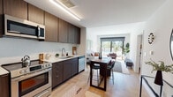 Located at 101 Manchester Rd in Richmond, VA, this modern single-floor residence offers a thoughtfully designed open-concept layout that seamlessly connects the kitchen, dining, and living areas. The kitchen is outfitted with dark wood cabinetry, stainless steel appliances including a refrigerator, range, and microwave, and clean white countertops that provide ample prep space. Light hardwood flooring flows throughout, enhancing the bright and airy atmosphere. A bar-height dining table bridges the kitchen and living area, ideal for casual dining or entertaining. The living room is anchored by large sliding glass doors that flood the space with natural light and open to an outdoor area with views of a pool and surrounding greenery, creating a seamless indoor-outdoor connection. The bedroom is a calm retreat featuring a ceiling fan, wide windows with slatted blinds, and warm wood-toned flooring. The bathroom maintains the home's modern aesthetic with large-format tile flooring, dark wood vanity cabinetry, and a full tub-shower combination. Clean white walls throughout lend a cohesive, contemporary feel to this well-designed Richmond residence.