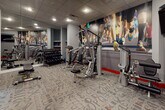 Located at 50 Barnum Ave in Port Washington, NY, this single-floor fitness center is designed to provide a comprehensive workout experience in a well-equipped, modern setting. The space features a bold interior design with grey walls accented by striking red baseboards, a patterned carpet floor with typographic motifs, and a drop-panel ceiling with recessed lighting throughout.

The cardio zone houses Matrix-brand treadmills and an elliptical trainer, positioned near a narrow horizontal window that allows a glimpse of natural light and outdoor greenery. The strength training area is well-stocked with a variety of resistance machines, including a cable system and selectorized equipment, complemented by a full rack of dumbbells displayed along a mirrored wall that enhances the sense of depth and space.

A large, vibrant mural of marathon runners serves as a dynamic focal point, energizing the atmosphere and reinforcing the facility's athletic spirit. A wall-mounted flat-screen television is positioned for visibility across the workout floor. The space also includes a water dispenser station, sanitation supplies, and a clearly marked emergency exit, ensuring both convenience and safety for users. The overall ambiance is functional, motivating, and thoughtfully appointed for a well-rounded fitness experience.