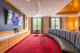 This single-floor amenity space is thoughtfully designed to offer a variety of social and functional zones within one cohesive environment. The main lounge area serves as the centerpiece, anchored by a sweeping curved wooden sofa with striped upholstery that faces a large wall-mounted flat-screen television set against a teal blue shiplap accent wall. A streamlined wooden media console with paneled cabinetry runs beneath the screen, adding warmth and organization to the space. Recessed lighting and crown molding along the white ceiling lend a polished, refined character throughout.

Bold, red patterned carpeting with a circular motif unifies the entire space and adds a striking visual energy underfoot. Large windows dressed with floor-length drapes allow ample natural light and views of surrounding greenery. A quieter seating nook features a sleek linear fireplace framed in stone tile, flanked by contemporary wall sconces and modern lounge chairs, creating a sophisticated retreat.

A compact meeting room sits off the main area, furnished with a round dark table, four chairs, a wall-mounted television, and colorful artwork, making it ideal for small gatherings. A decorative console area with pendant lighting and mirrored wall accents completes the space, reinforcing its polished, hospitality-inspired design.