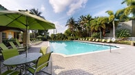 This inviting outdoor amenity space is defined by its generously sized swimming pool, set against a backdrop of towering palm trees and dense tropical greenery that creates a resort-like atmosphere. The pool deck is finished with distinctive hexagonal paver tiles that extend across the entire outdoor area, providing both visual texture and a durable, slip-resistant surface. Ample poolside seating is arranged throughout, including lime-green lounge chairs and umbrella-shaded dining sets that offer comfortable spots for sunbathing or casual gatherings. An arched, covered patio area adjoins the main building, featuring ceiling fans for added comfort and providing a shaded transition zone between the interior and the pool deck. The covered patio is enclosed by warm yellow stucco walls accented with white-framed French doors and multi-pane windows, allowing natural light to flow through while maintaining a visual connection to the pool area. The architecture reflects a Florida-inspired Mediterranean style, with archways and stucco finishes that complement the lush outdoor setting. The combination of open sky, swaying palms, and well-appointed outdoor furniture creates an atmosphere that is both relaxing and functional, making this space well-suited for leisure and social activities alike.