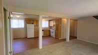This two-floor space offers a practical and open layout on its main level, where the kitchen and living area flow seamlessly together. The kitchen is well-equipped with white appliances, including a full-size refrigerator and a gas range, complemented by an abundance of warm wood-toned cabinetry that provides generous storage. A sink is positioned beneath a window dressed with white blinds, allowing natural light to filter into the space. The kitchen features hard surface flooring, which transitions into carpeting in the adjoining living area, creating a subtle distinction between the two zones. Structural columns are integrated into the open floor plan, adding architectural definition without fully separating the spaces. The textured popcorn ceiling runs throughout the main level, and recessed and flush-mount lighting provides functional illumination. The neutral color palette of warm beiges and tans throughout the walls, cabinetry, and flooring creates a cohesive and understated aesthetic. With two floors, the space offers additional living or sleeping areas beyond the main level.