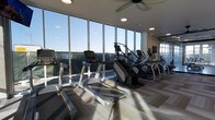 This well-appointed fitness center offers a bright, energizing environment designed to support a comprehensive workout experience. The space is defined by a striking wall of floor-to-ceiling windows that floods the interior with natural light and provides expansive outdoor views, creating an open and motivating atmosphere for exercise. The large-pane glazing gives the room an airy, elevated feel, making workouts feel less confined and more connected to the outdoors.

The cardio zone is prominently featured and well-equipped, housing multiple treadmills, stair climbers, and elliptical machines — all appearing to be modern, commercial-grade units with individual display consoles. The equipment is arranged in a thoughtful layout that maximizes use of space while allowing comfortable circulation between machines.

Underfoot, the flooring features a neutral, textured carpet tile that adds subtle warmth to the otherwise clean and contemporary aesthetic. Recessed ceiling lighting supplements the natural light, ensuring consistent illumination throughout the day. A ceiling fan overhead further enhances comfort during intense workouts.

The overall design is clean, modern, and functional, with white walls and a minimal color palette that keeps the focus on the equipment and the views. This fitness center delivers both form and function in a polished, professional setting.