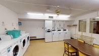 The Yellowwood laundry facility is a well-equipped, single-floor shared space designed to serve the needs of its users with practicality and efficiency. The laundry area is the centerpiece of the space, featuring multiple front-loading washers and top-loading dryers arranged in rows along the walls. Operating instructions are clearly posted, and utility hooks, shelving, and a laundry cart are available for convenience. A utility sink provides additional functionality in a dedicated alcove, and baseboard heating ensures year-round comfort.

Adjacent to the laundry area is a common sitting area with a distinctive curved wall design, lined with a row of interior-facing windows that look out into a neighboring hallway or common area, creating a sense of openness. A bulletin board displays notices and informational materials, while a wall clock keeps track of time. A folding table with chairs provides a comfortable place to wait. A vending machine near the entrance offers added convenience for users.

Throughout the space, the flooring is a neutral, easy-to-clean vinyl, and the textured white ceilings are fitted with fluorescent lighting that keeps the area bright and functional. The overall layout is clean, organized, and straightforward, making this a reliable and accessible shared amenity.