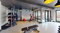 Located at 8108 Grace Farms Dr. in Charlotte, NC, this single-floor fitness center is thoughtfully designed to accommodate a full spectrum of workout needs. The space is organized into distinct zones, each purpose-built for a different type of training. The main cardio area features a long row of treadmills, ellipticals, and stationary bikes, all positioned along large windows that let in abundant natural light and offer views of the surrounding outdoor areas. The strength training section is outfitted with cable machines, multi-functional racks, adjustable benches, and a comprehensive dumbbell rack, complemented by a wall-mounted flat-screen TV. A dedicated functional fitness studio features warm hardwood flooring and is equipped with battle ropes, TRX suspension trainers, kettlebells, medicine balls, and resistance bands organized on wall-mounted storage racks. Glass partition walls and sliding doors separate zones while maintaining visual openness throughout. The design aesthetic is modern industrial, with dark tray ceilings contrasting crisp white walls, and bold black pendant lights with gold interiors providing focused illumination. Large ceiling fans and recessed lighting ensure the space remains well-ventilated and bright. Outdoor access through glass doors connects the facility to an exterior terrace.