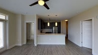This single-floor residence features an open-concept layout that seamlessly integrates the kitchen and living area. The kitchen is outfitted with dark espresso cabinetry, a mosaic tile backsplash, and stainless steel appliances, including a full-size refrigerator. A peninsula with a light-toned countertop extends into the living space, providing both additional prep area and casual seating. Two pendant lights hang above the peninsula, complementing the overall modern aesthetic.

The living area is equipped with a ceiling fan with integrated lighting, adding both function and style to the space. Wood-look plank flooring runs throughout, lending warmth and continuity across the open floor plan. Neutral-toned walls create a clean, versatile backdrop that suits a variety of furnishing styles.

A doorway visible from the main living area leads to an adjacent room, suggesting a bedroom or hallway beyond, while an additional passage hints at further living spaces. The layout feels open and well-connected, with consistent finishes tying each area together cohesively. The overall ambiance is contemporary and move-in ready.