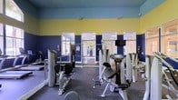 This fitness center is a single-floor facility designed to provide a comprehensive and functional workout environment. The space is immediately distinguished by its bold two-tone wall design, featuring deep navy blue on the lower half and a vibrant yellow on the upper portion, creating an energetic and motivating atmosphere. The layout is open and spacious, allowing for comfortable movement between equipment stations. The facility is outfitted with professional-grade Hoist strength training machines, including leg press, seated row, and various resistance machines strategically arranged throughout the floor. Large mirrored panels line the walls, enhancing the sense of space and allowing users to monitor their form during workouts. A mounted flat-screen television is positioned prominently on the wall, adding to the amenity-rich environment. The flooring consists of durable dark rubber or carpet-style material, well-suited for a high-traffic fitness setting. Natural light filters in through multiple windows and glass-paneled doors, brightening the interior and connecting the space to its surroundings. Overhead lighting ensures consistent illumination throughout. The overall ambiance is clean, modern, and purpose-built, making this fitness center a well-appointed facility for strength training and general exercise.