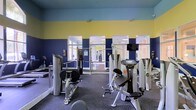 This fitness center is a single-floor facility designed to provide a comprehensive and functional workout environment. The space is immediately distinguished by its bold two-tone wall design, featuring deep navy blue on the lower half and a vibrant yellow on the upper portion, creating an energetic and motivating atmosphere. The layout is open and spacious, allowing for comfortable movement between equipment stations. The facility is outfitted with professional-grade Hoist strength training machines, including leg press, seated row, and various resistance machines strategically arranged throughout the floor. Large mirrored panels line the walls, enhancing the sense of space and allowing users to monitor their form during workouts. A mounted flat-screen television is positioned prominently on the wall, adding to the amenity-rich environment. The flooring consists of durable dark rubber or carpet-style material, well-suited for a high-traffic fitness setting. Natural light filters in through multiple windows and glass-paneled doors, brightening the interior and connecting the space to its surroundings. Overhead lighting ensures consistent illumination throughout. The overall ambiance is clean, modern, and purpose-built, making this fitness center a well-appointed facility for strength training and general exercise.