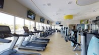 This fitness center is thoughtfully designed to support a comprehensive workout experience. The space features an open, single-floor layout with light hardwood flooring that creates a clean, energetic atmosphere throughout. Large floor-to-ceiling windows line one wall, flooding the interior with natural light and providing views of the surrounding outdoor environment, enhancing the overall workout experience.

The cardio zone is prominently featured, with a row of Matrix treadmills and elliptical trainers arranged along the windowed wall, each equipped with individual screens for entertainment. Multiple wall-mounted flat-screen televisions are positioned throughout the space for convenient viewing from various workout stations. Ceiling fans help maintain comfortable airflow across the open floor plan.

Toward the rear of the space, a dedicated strength training area houses a selection of Hoist resistance machines and free weight equipment, supported by large mirrors that visually expand the room. A distinctive yellow pendant light fixture adds a bold design accent, complementing the neutral cream-toned walls and bright, modern aesthetic of the facility overall.