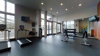This fitness center is designed with both functionality and comfort in mind, offering a spacious, open layout finished with durable dark rubber flooring suited for a variety of workout routines. The space features impressively high ceilings that contribute to an airy, open atmosphere, enhanced further by floor-to-ceiling glass doors and tall windows along the exterior wall, flooding the interior with abundant natural light and providing views of the surrounding area.

The facility is well-appointed with a range of equipment to support diverse fitness needs. Cardio options include a treadmill and a stationary bike equipped with a digital display console. On the strength training side, a multi-tiered dumbbell rack holds a comprehensive selection of free weights, with additional dumbbells accessible on the floor nearby. A wall-mounted flat-screen television is positioned for easy viewing during workouts, and a full-length mirror along one wall enhances the sense of space while allowing users to monitor their form. A water fountain and waste receptacle are conveniently located near the entrance, completing the practical, user-focused design of this well-maintained facility.