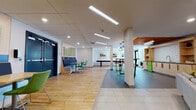This well-appointed single-floor communal space is designed to offer a versatile and inviting environment for social gathering and leisure. The open-plan layout seamlessly connects several distinct functional zones, each thoughtfully designed with its own character and purpose.

The main lounge area features a combination of large-format light tile and warm wood-plank flooring, creating a visual distinction between zones while maintaining a cohesive aesthetic. Recessed and linear LED lighting throughout the drop-panel ceiling ensures consistent, bright illumination across the entire space.

A dedicated seating nook lines one wall with continuous navy blue upholstered bench seating, complemented by individual wood-top tables paired with vibrant green chairs. Decorative green acoustic panels are mounted above, adding both texture and sound absorption to the area.

Adjacent to this is a casual media lounge with a grey sofa, a wall-mounted flat-screen television, and access through bold navy double doors. A game area is anchored by a classic shuffleboard table set against a wall displaying a series of framed urban photography prints.

Toward the far end of the space, a café-style bar area features high-top tables with metal stools, warm wood cabinetry with built-in shelving, a distinctive warm-toned wood slat ceiling accent, and pendant lighting, rounding out the space with an upscale, welcoming atmosphere.