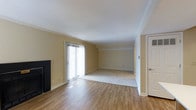 This single-floor residential space offers a well-designed open layout that blends comfort and functionality. The main living area is a standout feature, combining hardwood flooring in the primary zone with plush carpeting in an adjoining space, creating a natural distinction between areas while maintaining an open, connected feel. Crown molding runs along the ceiling, adding a refined architectural detail that elevates the overall aesthetic of the room. A fireplace anchors one side of the living area, providing a classic focal point and a cozy atmosphere. Large sliding glass doors dressed with vertical blinds allow abundant natural light to pour into the space, brightening the warm, neutral-toned walls and creating an airy, inviting ambiance. The doors also provide direct access to an outdoor area, seamlessly connecting the interior to the exterior. The ceiling height contributes to the sense of openness throughout. Clean white trim and baseboards complement the neutral color palette, giving the space a timeless and versatile character. The layout offers a practical flow, making the living area suitable for a variety of arrangements and uses. Overall, this space presents a bright, classic residential environment with thoughtful design details throughout.