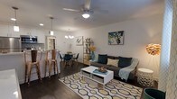 This well-appointed single-floor residence showcases a thoughtfully designed open-concept layout that seamlessly connects the living, dining, and kitchen areas, creating a cohesive and inviting atmosphere. The living area is anchored by a comfortable gray sofa accented with teal and patterned throw pillows, centered around a modern white coffee table resting on a bold geometric area rug. Rich dark hardwood flooring runs throughout, adding warmth and contrast to the bright white walls. Colorful contemporary artwork adds personality and visual interest to the space. A ceiling fan with integrated lighting provides both comfort and illumination overhead. The dining area features a round table surrounded by eclectic mixed seating, lit by a stylish pendant chandelier. Adjacent to the dining space, a kitchen bar counter lined with copper-toned metal stools offers additional casual seating and connects effortlessly to the kitchen beyond, which features tiled backsplash detailing visible in the background. Pendant lighting fixtures are placed thoughtfully throughout to enhance the ambiance. An open metal shelving unit serves as a functional and decorative element, bridging the living and dining zones. The overall aesthetic is modern, warm, and eclectic, making this a comfortable and visually dynamic living environment.
