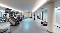 Located at 703 E Main St in Richmond, Virginia, this contemporary fitness center offers a comprehensive workout environment spread across a single, expansive floor. The space is designed with a clean, modern aesthetic, featuring smooth light-gray flooring, crisp white walls, and a sophisticated recessed lighting system that runs along the ceiling perimeter, creating a bright and energizing atmosphere throughout.

The facility is organized into distinct functional zones. A dedicated cardio area lines one side of the main floor, housing a row of treadmills, stair climbers, and elliptical machines, each equipped with individual display screens. Adjacent to this zone, a strength and free-weight area features a well-stocked rack of dumbbells, kettlebells, resistance bands, and weight plates alongside a power rack for barbell training. A separate cycling studio accommodates multiple stationary spin bikes, reflected in floor-to-ceiling mirrors that enhance the sense of space. Adjustable weight benches and functional training sleds round out the equipment offerings.

Large mirrors are strategically placed throughout, amplifying the natural and ambient light while making the space feel open and expansive. Black-framed glass doors and a skylight add architectural interest and allow additional light to filter in. The overall ambiance is clean, professional, and well-organized, making it a fully equipped destination for a wide range of fitness activities.