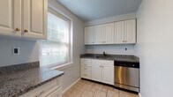 This single-floor residence offers a thoughtfully designed layout with a clean, neutral aesthetic throughout. The kitchen is a functional highlight, featuring white raised-panel cabinetry with gold hardware, dark granite countertops, a classic white tile backsplash, a stainless steel dishwasher, and light stone-tiled flooring. Large windows with blinds allow generous natural light to fill the space, creating an airy and welcoming atmosphere.

The living areas and bedrooms are finished with rich, warm-toned hardwood flooring that runs throughout. Multiple rooms benefit from natural light via double-hung windows with views of surrounding greenery. Classic radiator units provide supplemental heating in the living and bedroom areas, while a wall-mounted mini-split unit offers additional climate control. The rooms feature soft gray walls and clean white baseboards, contributing to a calm, cohesive interior palette.

The bathroom is finished with white subway-style wall tiles, dark floor tiles, and a soaking tub with chrome fixtures, complemented by a window for natural ventilation. A dedicated laundry area houses a stacked washer and dryer unit for added convenience. The entry hallway provides a welcoming transition into the home, completing this well-equipped and comfortable single-floor living space.