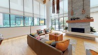 This contemporary single-floor communal space is defined by its dramatic double-height ceilings that lend an impressive sense of openness and grandeur throughout. Warm-toned hardwood flooring runs seamlessly across the entire space, unifying distinct functional zones into a cohesive and inviting environment.

The main lounge area features a generous sectional sofa and mid-century modern accent chairs arranged around a solid wood cube coffee table, all anchored by a patterned area rug. A striking stacked-stone fireplace serves as a focal point, flanked by lush potted plants and decorative oars. Oversized floor-to-ceiling windows and glass doors bathe the space in abundant natural light while offering direct views to an outdoor terrace.

A secondary seating zone is elegantly appointed with upholstered armchairs, a central game table, and a wall-mounted flat-screen TV, with clusters of sleek cylindrical pendant lights descending dramatically from above. Colorful artwork adds visual interest to the crisp white walls.

A café-style dining and lounge area features round white tables paired with teal accent chairs alongside a raised bar counter lined with sculptural walnut barstools. Wooden slatted partition screens define separate areas while maintaining an open, airy feel. The overall ambiance is polished, welcoming, and thoughtfully designed for both relaxation and social interaction.