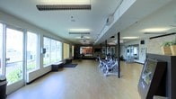 This single-floor fitness center offers a clean, functional layout designed to support a comprehensive workout experience. The space is anchored by gleaming hardwood-style flooring that spans the entire area, providing a polished and durable surface for a variety of exercise activities. A row of stationary cycling bikes lines one side of the room, positioned for group or individual use, while a dedicated strength training and cable machine station is visible along the opposite wall. Full-length mirrors extend across a large section of the wall, enhancing the sense of depth and allowing users to monitor their form during workouts. Natural light pours in through a generous bank of large windows along one side, offering views of a well-maintained outdoor landscape. Ceiling fans are mounted overhead to ensure comfortable airflow throughout the room. Fluorescent lighting provides consistent, even illumination throughout. A dark area rug near the entrance adds a subtle design touch and serves as a transitional zone into the workout area. The overall ambiance is bright, open, and purposefully organized, making the space welcoming and practical for a wide range of fitness needs.