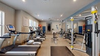 This two-floor exercise facility in Houston's 77003 district offers a comprehensive fitness environment spread across two well-appointed levels, connected by a staircase with an elegant dark wood and wrought iron railing. The ground floor serves as the primary gym space, featuring a generously sized workout area with plush carpeted flooring and ample natural light from multiple windows. It is outfitted with a wide array of cardio machines, including treadmills and ellipticals, alongside a full rack of dumbbells, a cable weight machine, a weight bench, and additional strength training equipment. Floor-to-ceiling mirrors line one wall, enhancing the sense of space, while multiple ceiling fans maintain airflow throughout. A large flat-screen TV is mounted for entertainment during workouts. The upper level is dedicated to cycling and open exercise, featuring light hardwood flooring that gives the space a bright, airy feel. Spinning bikes are arranged facing a mirrored wall, and dual ceiling fans keep the room comfortable. A wall-mounted TV and decorative artwork complete the space. Recessed lighting throughout both floors ensures a well-lit, welcoming atmosphere for all types of workouts. Together, the two floors create a versatile and fully equipped fitness center suited for a wide range of exercise routines.