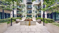 This open-air courtyard in New Orleans, LA, is a well-designed outdoor space nestled within a multi-story building complex. The courtyard's architecture is characterized by clean, contemporary lines, with the surrounding structure featuring large steel-framed grid windows, smooth light-colored stucco facades, and stacked balconies with horizontal metal railings that overlook the shared space below.

At the heart of the courtyard sits a raised water feature with a shallow basin, flanked by a pair of wooden benches that provide a tranquil seating area. Surrounding this central focal point are generously sized concrete planters filled with manicured shrubs and slender deciduous trees, whose canopies introduce natural shade and seasonal greenery. Additional planted beds line the perimeter, reinforcing the courtyard's structured yet inviting landscape design.

The ground level is finished with large-format stone pavers that extend throughout, creating a clean, cohesive surface. Ground-floor units open directly onto the courtyard through steel-framed glass doors and feature private patio areas enclosed by horizontal metal railings, some furnished with outdoor seating. The open-sky orientation ensures ample natural light throughout the day, making this courtyard a functional and visually appealing communal outdoor environment.