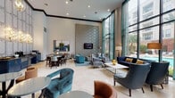 The Social Lounge in Houston, TX (77057) is a thoughtfully designed communal space that blends contemporary elegance with comfortable functionality across a single, expansive floor. Dramatic double-height ceilings give the lounge a grand, airy atmosphere, while floor-to-ceiling windows framed by teal drapery flood the interior with natural light and offer sweeping views of a resort-style pool and surrounding architecture.

The space is organized into several distinct seating zones. A primary lounge area features a tufted navy blue sofa, patterned accent chairs, and a sleek white coffee table positioned atop a teal area rug, all set against a boldly textured, patterned feature wall with a wall-mounted flat-screen television. Adjacent to this is a more intimate conversation area with teal and cognac barrel chairs arranged around glass-topped tables beneath gold crystal pendant chandeliers, accented by curated wall art.

A dining and refreshment area is anchored by a round table with upholstered chairs and is complemented by navy cabinetry with dark countertops, built-in appliances, and an ornate paneled accent wall illuminated by gold sconce lighting. Light-toned marble flooring flows throughout, unifying the various zones into a cohesive, sophisticated environment ideal for socializing and entertaining.