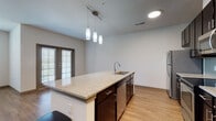 This well-designed single-floor residence offers a thoughtfully planned layout that balances open communal areas with comfortable private spaces. The kitchen serves as the centerpiece, featuring a large granite-topped island with an integrated sink, dark espresso cabinetry, a subway tile backsplash, and a full suite of stainless steel appliances including a refrigerator, range, and microwave. Pendant lighting hangs above the island, providing both ambient and task illumination. The kitchen flows seamlessly into the living area, which is finished with warm hardwood-style flooring and benefits from natural light through sliding glass doors leading to an outdoor access point. The bedrooms are generously proportioned, with ceiling fans, plush carpeting or hardwood-style flooring depending on the room, and large windows fitted with blinds that invite natural light while maintaining privacy. One bedroom also features built-in shelving, adding practical storage. The bathroom is appointed with a granite vanity, a soaking tub with tile surround, and direct access to a dedicated closet space. Throughout the residence, consistent finishes including warm wood tones, neutral walls, and modern fixtures create a cohesive and inviting atmosphere.
