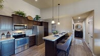 This single-story residence is thoughtfully designed with a cohesive neutral color palette and wood-look flooring that flows throughout the main living areas. The kitchen serves as a central highlight, showcasing rich dark wood cabinetry that extends to the upper walls, a stone tile backsplash, light granite-style countertops, and a full suite of stainless steel appliances including a refrigerator, range, microwave, and dishwasher. Soaring ceilings above the kitchen add a sense of openness. An elongated island with bar-height seating accommodates multiple guests and is illuminated by two elegant pendant lights, making it ideal for dining or entertaining. The living area offers a comfortable, intimate layout with neutral-toned walls and ample room for seating arrangements. Multiple bedrooms feature plush carpeting, ceiling fans with light fixtures, and windows that allow natural light. The bathroom includes a dark vanity with granite-style countertops, a wide mirror with a multi-bulb light fixture, and a tub-shower combination. A dedicated laundry area provides overhead dark wood cabinetry and a wall-mounted drying rack for added convenience.
