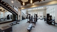 This two-story fitness center is designed to support a full range of workout routines within a thoughtfully organized, modern facility. The main floor is anchored by soaring double-height ceilings that lend the space a sense of openness, further enhanced by rows of tall windows along multiple walls that allow generous natural light to pour in and provide views of the surrounding outdoor amenity areas, including what appears to be a pool deck. The floor is finished in neutral carpet tile and equipped with an extensive selection of strength machines, cable systems, free weights, benches, and cardio equipment including multiple treadmills and stationary bikes. A starburst pendant fixture adds a decorative focal point overhead, complementing the recessed lighting throughout. A dedicated entry area features a built-in storage unit for towels and accessories, alongside full-length mirrors and stability balls for functional training. An industrial-style open staircase leads to the second floor, which houses a dedicated cycling studio finished with light hardwood flooring, floor-to-ceiling mirrors, a large ceiling fan, and a wall of windows. A separate room on the upper level includes a large-screen display, mirrors, and direct outdoor access, providing a versatile space for group fitness or personal training. The overall design balances functionality with a clean, contemporary aesthetic.