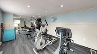 This well-appointed fitness center in Spokane, WA offers a comprehensive workout environment designed to support a variety of exercise routines. The space is finished with durable carpet-tile flooring and features recessed lighting throughout, creating a clean and functional atmosphere. Soft blue accent walls, wainscoting trim, and motivational wall decals — including "DOnt quIT" and "MAKE MUSCLES" — contribute to an energizing and encouraging ambiance. A wall of mirrors along one side visually expands the room and aids in form correction during workouts. Cardio equipment includes True-brand upright and recumbent stationary bikes, elliptical trainers, and additional cardio machines, while a multi-station cable strength machine and free weight area with stability balls and a flat bench round out the strength training options. A wall-mounted flat-screen television provides entertainment during workouts. Natural light flows in through multiple windows and glass doors that open to an outdoor view, keeping the space feeling open and airy. The single-floor layout ensures easy accessibility throughout, making this fitness center a practical and well-equipped amenity for its users.
