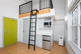 Located in Seattle's Capitol Hill neighborhood, this modern loft-style unit at 1815 Bellevue Avenue offers a thoughtfully designed, single-floor layout with striking architectural details. The main living area is defined by soaring double-height ceilings and an expansive wall of floor-to-ceiling windows that flood the space with abundant natural light and frame views of the surrounding Seattle streetscape. Warm hardwood-style flooring runs throughout, complementing the clean, neutral-toned walls.

A distinctive built-in loft level is accessed via a sleek steel ladder, providing an elevated secondary space ideal for sleeping or storage. Beneath the loft, a compact kitchenette is efficiently integrated with white cabinetry, a microwave, and a mini refrigerator. A vibrant chartreuse entry door adds a bold pop of color and serves as a memorable architectural accent.

Large sliding glass doors open directly onto a spacious private deck, finished with light gray composite decking and framed by black steel railings. The deck offers open-air views of the surrounding Capitol Hill neighborhood and receives ample sunlight throughout the day. A frosted glass window provides additional natural light to an adjacent area while maintaining privacy. This unit combines contemporary urban design with functional, efficient living in a well-connected Seattle location.