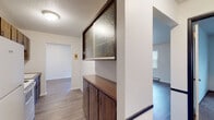 This single-floor unit offers a straightforward, functional layout designed for comfortable everyday living. The entryway features a distinctive built-in storage unit with warm-toned wood cabinetry below and a framed panel of textured, frosted glass above, adding a subtle decorative element that separates the entry from adjoining spaces.

The galley-style kitchen is equipped with wood-toned upper and lower cabinetry, a double-basin sink, an electric range, and a full-size refrigerator. Light wood-look flooring runs underfoot, and a textured ceiling ties the space together. An adjacent pass-through area leads to additional storage.

The living area is generously proportioned, featuring dark wood-look flooring, multiple windows that allow natural light to fill the room, a wall-mounted air conditioning unit, and a baseboard heater along the exterior wall. A secondary living or dining area with lighter flooring and its own baseboard heater provides added flexibility.

The bedroom is carpeted and bright, with a window fitted with blinds and a baseboard heater beneath it, creating a cozy atmosphere. The bathroom includes a wide vanity with a tan countertop, a framed mirror, and wood-look flooring, offering a clean and practical design throughout.