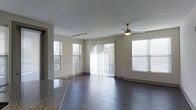 This single-floor residence offers a well-designed open-concept layout that seamlessly connects the living and kitchen areas. The living space is anchored by rich, dark hardwood flooring that extends throughout, providing a sleek and modern aesthetic. Neutral-toned walls create a clean, versatile backdrop that complements the overall design.

Natural light floods the interior through multiple windows fitted with horizontal blinds, as well as a full-length sliding glass door dressed with vertical blinds, offering both brightness and privacy control. The sliding door provides convenient access to an outdoor area, framing views of surrounding greenery.

Overhead, a ceiling fan with an integrated light fixture serves the living area, ensuring comfort year-round, while a track lighting system in the adjacent kitchen zone adds focused, adjustable illumination. A portion of a granite countertop is visible at the edge of the frame, hinting at quality finishes in the kitchen area.

The open layout and generous window placement give the space an airy, expansive feel. The combination of practical features — from the durable flooring to the thoughtful lighting design — makes this a functional and inviting living environment suited for everyday comfort.