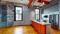 This single-floor loft space masterfully combines industrial heritage with contemporary design. Exposed brick walls and bold red structural steel beams run throughout, creating a striking visual identity that honors the building's historic character. Warm hardwood floors extend across the entire space, adding natural richness to the raw aesthetic.

The kitchen is a standout feature, equipped with stainless steel appliances including a gas range and built-in microwave, granite countertops, a deep undermount sink with a matte black faucet, and a bold orange island that serves as a vibrant focal point. White shaker-style cabinetry lines the kitchen walls, balancing the industrial elements with clean, modern detailing. Large black-framed windows flood the kitchen with natural light while offering views of the surrounding urban streetscape.

A long, well-lit corridor connects the various rooms, with exposed ductwork and track lighting overhead maintaining the industrial theme. The open living areas benefit from high ceilings, ceiling fans, and additional natural light from generously sized windows set into the original brick walls.

The bathroom features marble-tiled walls, a glass-enclosed shower, a soaking tub, and granite vanity countertops with matte black fixtures, delivering a refined, spa-like finish that contrasts beautifully with the space's raw industrial backbone.