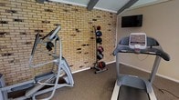 This single-story fitness space offers a well-equipped workout environment characterized by its distinctive exposed brick wall, which serves as a striking architectural backdrop. The ceiling features visible structural beams, adding a raw, industrial character to the room. Durable, low-pile carpet covers the floor, providing adequate cushioning for exercise activities.

The room is outfitted with key cardio equipment, including an elliptical trainer and a treadmill, both positioned to make efficient use of the available floor space. A vertical medicine ball rack holds multiple weighted balls of varying sizes, offering strength and conditioning options alongside the cardio machines. A flat-screen television is mounted on the wall, positioned at a comfortable viewing angle from the equipment, enabling users to watch content during workouts.

The space is compact yet functional, designed to accommodate essential fitness activities without excess. The combination of brick walls, exposed beams, and practical equipment creates an atmosphere that is both utilitarian and characterful, making it a straightforward and purpose-driven exercise environment.