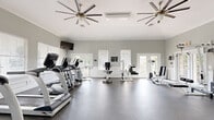 Located at 100 Riverchase Boulevard in Nashville, TN, this well-equipped fitness center offers a spacious, open-concept layout designed to support a full range of workout routines. The interior is finished with a neutral gray color palette, crisp white crown molding, and white wainscoting that lends a clean, polished aesthetic throughout.

The flooring consists of durable speckled rubber, ideal for both cardio and strength training activities. Two large ceiling fans with brushed nickel finishes ensure consistent airflow across the workout floor. Multiple wall-mounted flat-screen televisions are positioned for easy viewing from various exercise stations.

The cardio section features a row of treadmills and additional aerobic equipment lined along one wall, while the strength training area includes a cable machine, power rack, adjustable benches, and a full dumbbell rack. Multiple windows fitted with horizontal blinds allow abundant natural light to fill the space, while French doors on multiple walls provide views of the surrounding greenery and convenient access to the outdoors.

The open floor plan allows for generous spacing between equipment, promoting a comfortable and unobstructed workout experience. Overall, this fitness center combines functional design with an inviting atmosphere, offering a comprehensive exercise facility well-suited for a variety of fitness goals.