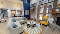 Located at 4001 W Northgate Dr in Irving, Texas, this single-floor community clubhouse showcases a thoughtfully designed interior that balances bold aesthetics with comfortable, functional spaces. The main lounge area immediately draws attention with its soaring vaulted ceilings and striking cobalt blue accent walls, creating a dramatic yet inviting atmosphere. Large multi-pane windows dressed with patterned draping panels line the walls, flooding the interior with abundant natural light and offering views of the surrounding outdoor greenery.

The lounge is anchored by a grand floor-to-ceiling stacked stone fireplace with a wood mantel, flanked by vivid yellow wingback chairs and complemented by a sectional sofa arranged over a chevron-patterned area rug. Wood-look flooring extends throughout, adding warmth and continuity. A sputnik-style chandelier adds a mid-century modern touch overhead.

Adjacent to the lounge, exposed brick walls frame a curved bar-style counter finished in dark granite with seating for several guests, ideal for social gatherings. A dining area nearby features a mounted television and ample seating. An open staircase with brick detailing leads to additional areas, while a business or leasing office is visible in the background, completing this well-appointed communal space.
