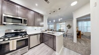 This single-floor residence is designed with a modern, cohesive aesthetic that combines functionality and style across every room. The heart of the home is an open-concept kitchen, equipped with rich dark-stained cabinetry, crisp white quartz countertops, a subway tile backsplash, and a full suite of stainless steel appliances including a gas range, built-in microwave, and dishwasher. A peninsula with an undermount sink and pendant lighting above connects the kitchen seamlessly to the dining and living areas, creating an inviting, social layout. Light wood-look flooring runs through the main living areas, transitioning to plush carpet in the living room, dining room, and bedroom. The living room features crown molding, generous natural light from large windows dressed with sheer curtains, and a dedicated nook ideal for a home office setup. The dining room is framed by crown molding and offers ample space for a full dining set. The bedroom is a calm retreat, carpeted and naturally lit, with room for a king-sized bed. The bathroom includes a dark-stained vanity with white countertop and a shower-tub combination. A dedicated in-unit laundry room with a full-sized washer and dryer completes this thoughtfully designed residence.