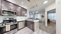 This single-floor residence is designed with a modern, cohesive aesthetic that combines functionality and style across every room. The heart of the home is an open-concept kitchen, equipped with rich dark-stained cabinetry, crisp white quartz countertops, a subway tile backsplash, and a full suite of stainless steel appliances including a gas range, built-in microwave, and dishwasher. A peninsula with an undermount sink and pendant lighting above connects the kitchen seamlessly to the dining and living areas, creating an inviting, social layout. Light wood-look flooring runs through the main living areas, transitioning to plush carpet in the living room, dining room, and bedroom. The living room features crown molding, generous natural light from large windows dressed with sheer curtains, and a dedicated nook ideal for a home office setup. The dining room is framed by crown molding and offers ample space for a full dining set. The bedroom is a calm retreat, carpeted and naturally lit, with room for a king-sized bed. The bathroom includes a dark-stained vanity with white countertop and a shower-tub combination. A dedicated in-unit laundry room with a full-sized washer and dryer completes this thoughtfully designed residence.