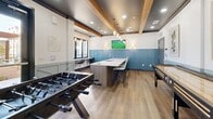 This amenity space is thoughtfully designed to offer both indoor and outdoor areas for recreation and socializing. The interior game room showcases a contemporary aesthetic, highlighted by exposed wood ceiling beams set against a dark ceiling, reclaimed-style hardwood flooring, and a striking blue subway tile accent wall that runs the length of the room. Pendant lighting and recessed fixtures create a warm, inviting atmosphere. The room is equipped with a foosball table and a shuffleboard table, while a central island with a sink and bar seating provides a convenient gathering spot. A wall-mounted flat-screen TV completes the entertainment setup, and a separate counter area along the window offers additional seating for more focused activities. The outdoor courtyard extends the social experience with a generous open-air layout framed by the surrounding multi-story building. A covered pergola structure with rich wood slatted privacy screens shelters picnic tables with pendant lighting overhead. Beyond the pergola, a natural gravel seating area is furnished with wooden lounge chairs and cylindrical accent tables, complemented by manicured greenery, raised planting beds, and string lights that lend a relaxed, communal ambiance ideal for gatherings throughout the day and evening.