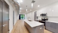 This contemporary single-floor residence showcases a thoughtfully designed open-concept layout with clean lines and modern finishes throughout. Light hardwood flooring flows seamlessly across the main living areas, complementing the bright, white walls and recessed lighting that give the space an airy, welcoming feel.

The kitchen is a standout feature, anchored by a large island with a white quartz countertop, an integrated sink with a sleek gooseneck faucet, and a built-in wine rack below. Two-tone cabinetry — gloss white uppers paired with matte gray lowers — surrounds a full suite of stainless steel appliances, including a refrigerator, range, and microwave. Exposed ductwork along the ceiling adds an industrial-chic character to the space.

The open living area extends from the kitchen toward a floor-to-ceiling sliding glass door that opens to a private balcony overlooking lush green trees, filling the room with natural light and outdoor views.

The bedroom is generously proportioned, featuring a bold gray accent wall, a ceiling fan, and a large black-framed window with serene tree views. An adjacent full bathroom is finished with large-format beige floor tiles, a dark-toned floating vanity with a white countertop, and a tub-shower combination with glass sliding doors.