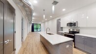 This contemporary single-floor residence showcases a thoughtfully designed open-concept layout with clean lines and modern finishes throughout. Light hardwood flooring flows seamlessly across the main living areas, complementing the bright, white walls and recessed lighting that give the space an airy, welcoming feel.

The kitchen is a standout feature, anchored by a large island with a white quartz countertop, an integrated sink with a sleek gooseneck faucet, and a built-in wine rack below. Two-tone cabinetry — gloss white uppers paired with matte gray lowers — surrounds a full suite of stainless steel appliances, including a refrigerator, range, and microwave. Exposed ductwork along the ceiling adds an industrial-chic character to the space.

The open living area extends from the kitchen toward a floor-to-ceiling sliding glass door that opens to a private balcony overlooking lush green trees, filling the room with natural light and outdoor views.

The bedroom is generously proportioned, featuring a bold gray accent wall, a ceiling fan, and a large black-framed window with serene tree views. An adjacent full bathroom is finished with large-format beige floor tiles, a dark-toned floating vanity with a white countertop, and a tub-shower combination with glass sliding doors.