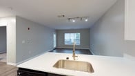 Unit 417 is a well-appointed 1-bedroom, 1-bathroom residence designed with a clean, modern aesthetic across a single floor. The open-concept living and kitchen area is anchored by durable wood-style flooring, neutral gray walls, and track lighting that illuminates the space effectively. A large window in the living area welcomes natural light and offers views of the surrounding greenery outside.

The kitchen is a standout feature, boasting a two-tone design with dark lower cabinets and crisp white upper cabinets, complemented by white countertops and a deep stainless steel undermount sink. Stainless steel appliances — including a gas range, over-the-range microwave, and full-size refrigerator — complete the functional and stylish cooking space. A kitchen island provides additional counter space and serves as a natural divider between the kitchen and living area.

The bedroom is generously sized with plush wall-to-wall carpeting, neutral tones, and a window that fills the room with natural light and outdoor views. The full bathroom features a tub/shower combination with white tile accents highlighted by navy blue mosaic tile detailing, wood-style flooring, and a vanity with a white countertop. Throughout the unit, clean lines and a cohesive neutral palette create a calm and inviting atmosphere.