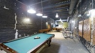 This single-floor space combines industrial design with recreational functionality. Exposed corrugated metal ceilings with visible ductwork and structural beams define the architectural character overhead, while dark painted brick walls lend a raw, urban aesthetic throughout. Pendant lights suspended from the ceiling provide focused illumination across the space.

The main area is anchored by a full-size pool table with a teal felt surface, positioned beneath large hanging industrial-style pendant lights. A shuffleboard table is visible along one wall, adding to the game room atmosphere. The flooring features a patterned carpet tile that grounds the space with subtle texture.

Toward the rear, a kitchenette area includes cabinetry and a countertop, alongside a wall-mounted flat-screen TV. A dining table with chairs and additional bar-height seating with metal stools offer multiple options for gathering. An accent wall in a warm green tone provides a visual contrast to the surrounding dark brick. Overall, the space delivers a casual, social atmosphere well-suited for entertainment and leisure.