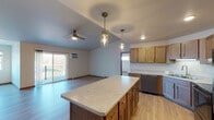This single-floor residence offers a thoughtfully designed open-concept layout, seamlessly connecting the kitchen and living areas for a functional and welcoming flow. The kitchen is a standout feature, equipped with warm-toned shaker-style cabinetry, light stone-patterned countertops, and a full suite of stainless steel appliances including an electric range with over-the-range microwave and a refrigerator. A central island with pendant lighting provides additional prep space and casual seating potential, while recessed lighting enhances the kitchen's bright, practical atmosphere. Light hardwood-style flooring runs throughout the main living spaces, complementing the neutral wall tones and creating a clean, cohesive aesthetic. The spacious living area benefits from natural light streaming through sliding glass doors that open to a private balcony, offering outdoor views of a natural landscape. A ceiling fan overhead adds comfort and airflow to the room. Baseboard heating is present throughout. The bathroom features light hardwood-style flooring, a marble-patterned vanity countertop, a soaking tub with shower, and a tall dark-toned storage cabinet for added organization. Interior doors with wood-toned finishes provide a consistent design element that ties the space together throughout.