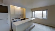 This single-floor unit offers a clean, open-concept layout that seamlessly connects the kitchen and living area. The kitchen is efficiently designed with abundant white upper cabinetry providing generous storage space. It is equipped with a white electric range and a built-in sink, all set within a compact, well-organized cooking zone. A white peninsula counter extends outward, creating a natural division between the kitchen and the adjoining living space while also offering additional prep or dining surface area. The living area features neutral wall-to-wall carpeting that contributes to a warm, understated ambiance. A large picture window serves as a focal point, flooding the space with natural light and framing views of neighboring buildings and tree-lined surroundings, suggesting an elevated position within the building. The neutral color palette throughout — soft whites and warm beiges — keeps the interior feeling bright and airy. The overall design prioritizes functionality and practicality, making efficient use of the available floor space. This unit is well-suited for those seeking a straightforward, low-maintenance living environment with comfortable access to natural light and urban views.