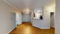 This single-floor residence at 1904 Parable Way in Cary, NC offers a well-organized layout with a cohesive design throughout. The main living area is generously proportioned, featuring warm hardwood-style flooring, crown molding, and soft gray walls that create a clean, neutral backdrop. A ceiling fan with a light fixture anchors the room, while multiple windows allow natural light to fill the space and provide views of the surrounding area.

The kitchen is positioned adjacent to the living area and is accessible via a peninsula with dark granite-style countertops that also functions as a breakfast bar. Warm-toned wooden cabinetry provides ample storage, complemented by matching granite counters, a stainless steel sink, and standard appliances including a refrigerator. The layout allows visual connectivity between the kitchen and living space.

The bedroom features plush carpeting and neutral walls, offering a quiet and comfortable retreat. The bathroom includes a full soaking tub and shower combination with white tile surround and chrome fixtures. A dedicated laundry area accommodates a full-size top-load washer with shelving above. A walk-in closet with wire shelving adds practical storage to the overall layout.